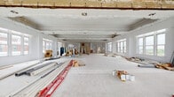 This single-floor space is a historically significant property currently in the midst of an active renovation. The building's architectural character is immediately evident through its elaborate, hand-crafted plaster moldings and ornamental detailing that run along the ceilings and upper walls throughout multiple rooms. These intricate decorative elements — including scrollwork, floral relief panels, and classical cornicing — speak to the building's heritage and craftsmanship.

The layout spans several generously proportioned, open rooms connected by wide doorways. Ceilings are notably high, lending an impressive sense of volume and grandeur to each area. Large, multi-pane windows line the exterior walls in nearly every room, flooding the interiors with natural light and framing views of the surrounding urban and natural landscape.

One area features a distinctive curved or rotunda-style alcove, adorned with an ornate carved plaster wall panel, adding further architectural interest. Structural columns are present in certain zones, contributing to the overall form of the space.

The floors are currently exposed concrete or bare subfloor throughout, awaiting finishes. While construction materials, scaffolding, and renovation supplies are presently visible, they reflect an ongoing transformation. This space presents a compelling opportunity to restore and repurpose a richly detailed historic interior with substantial square footage and outstanding natural light.