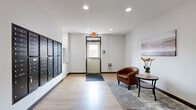 This residential building hallway serves as a functional and welcoming common area, thoughtfully designed to accommodate the daily needs of its residents. The space is finished with warm-toned luxury vinyl plank flooring throughout, complemented by clean white walls and dark wood baseboards that lend a modern, polished aesthetic.

At the core of the hallway is a dual-elevator bank, featuring sleek grey metal doors framed in dark trim, providing convenient vertical access throughout the building. Adjacent to the elevators, a secured stairwell door offers an additional access point. A prominent wall-mounted bank of dark metal mailboxes lines one side of the hallway, offering individual locked compartments for residents.

Near the main entry door, a cozy seating nook adds a residential touch, furnished with a tufted brown leather armchair, a round side table with a floral arrangement, and a soft area rug — creating an inviting spot to pause. A framed landscape artwork on the wall adds a decorative element to the space.

The main entry features a glass-paneled metal door, allowing natural light to filter into the hallway and providing a clear view to the exterior. Recessed ceiling lights and safety features including an emergency exit sign, fire extinguisher cabinet, and security camera ensure the space is both practical and compliant with safety standards.