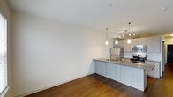 This single-floor space showcases a seamlessly integrated open-concept layout that connects the living area to a well-appointed kitchen. Rich, dark hardwood flooring runs throughout, providing a warm contrast to the crisp white walls and light-toned cabinetry. The kitchen is equipped with stainless steel appliances, including a refrigerator and range, complemented by a sleek tile backsplash and light stone countertops. A peninsula with a built-in sink and matching cabinetry defines the boundary between the kitchen and living area, offering both functional workspace and casual seating potential. Three stylish pendant lights hang above the peninsula, adding a contemporary focal point and targeted illumination. Additional track lighting over the kitchen workspace ensures thorough coverage for culinary tasks. The living area is open and versatile, with generous wall space and clean lines that invite flexible furniture arrangements. The overall ambiance is modern and understated, with neutral tones and quality materials contributing to a polished, move-in-ready aesthetic.