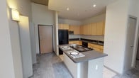 This single-floor loft-style unit showcases an industrial-modern design highlighted by exposed ductwork running along the ceilings, adding a distinct urban character throughout the space. The open-concept kitchen is a central feature, equipped with light maple cabinetry extending to the upper walls for generous storage, dark gray laminate countertops, and a suite of black appliances including a refrigerator, dishwasher, and built-in range. A peninsula island with an integrated cooktop provides additional prep space and casual seating, while tile flooring grounds the kitchen area cleanly. The main living area transitions seamlessly from the kitchen and features plush gray carpeting underfoot, bright white walls, and large windows that allow natural light to filter in. Recessed lighting in the kitchen and entry zone supplements the natural illumination throughout. The bedroom continues the light and airy aesthetic with soft carpet, crisp white walls, and a window offering views of the surrounding brick architecture. Exposed utility conduits on the ceiling reinforce the loft-inspired character of the space. The entry area includes a solid wood-core door and opens directly into the kitchen and living zones, creating an efficient and connected floor plan suited for modern urban living.