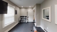 This dedicated fitness room offers a thoughtfully organized workout space finished with neutral gray walls, recessed lighting, and durable rubber-speckled flooring designed to withstand heavy equipment use. The room is laid out to maximize functionality, housing a variety of exercise equipment that supports both cardio and strength training routines.

On the cardio side, the space includes a treadmill and a NordicTrack elliptical machine, positioned to allow comfortable use without crowding. A stationary recumbent-style bike provides an additional low-impact option. A large flat-screen television is wall-mounted, positioned to be visible from multiple workout stations, making it easy to follow workout content or simply stay entertained during sessions.

For strength training, a tiered dumbbell rack holds a full range of free weights, neatly organized and easily accessible. A mirror panel on the wall aids in monitoring form. Natural light enters through windows fitted with white horizontal blinds, contributing to a bright and motivating atmosphere. Posted exercise room rules reflect a shared-use environment. The single-floor layout is practical and straightforward, with a standard door providing access to an adjacent space.
