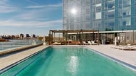 Located at 20 Hallets Point in Astoria, Queens, this expansive amenity floor offers a thoughtfully curated collection of spaces designed for wellness, work, and leisure. The entrance welcomes visitors with floor-to-ceiling glass doors, polished light-toned tile flooring, and a bright, airy corridor that sets an elegant tone throughout.

A dramatic curved wall clad in warm vertical wood slats serves as a striking architectural centerpiece, guiding residents through the floor with a sense of refined sophistication. Meeting and conferencing rooms are well-appointed, featuring round tables with vibrant orange chairs, wall-mounted displays, and generous natural light from nearby windows framing views of the waterfront and skyline.

Private meeting pods offer acoustically treated walls, textured geometric ceilings, and comfortable modern furnishings for focused work or collaboration. The screening lounge features tiered seating with plush light-grey sofas accented by navy and orange cushions beneath an ornate coffered ceiling.

The state-of-the-art fitness center is generously proportioned, equipped with cardio machines, free weights, and a mirrored strength training area, all beneath a tray ceiling with recessed lighting and warm wood accent walls.

Expansive floor-to-ceiling windows throughout frame sweeping views of the East River and the Manhattan skyline. Outdoor terraces extend the living space further, featuring lounge seating, landscaped planters, timber decking, and unobstructed panoramic city and waterfront views.