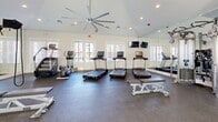 Located in Columbia, TN, this fitness center offers a comprehensive workout environment within a single open-plan floor. The space is designed with functionality and comfort in mind, featuring durable rubber flooring throughout that provides cushioning and traction during exercise. Recessed lighting and a large ceiling fan keep the room bright and well-ventilated, while multiple windows fitted with blinds allow natural light to fill the space without sacrificing privacy.

The cardio zone is prominently equipped with multiple treadmills and an elliptical machine, each featuring individual display screens for tracking workout metrics. Wall-mounted televisions provide entertainment options during cardio sessions. A full-length mirror along one wall visually expands the space and allows users to monitor their form.

The strength training area is equally well-appointed, featuring a multi-station cable machine, an adjustable weight bench, and a well-organized free weight rack stocked with a range of dumbbells and medicine balls. A cycling bike rounds out the cardio offerings for low-impact training options.

The clean, neutral color palette of soft white and grey walls contributes to a professional and motivating atmosphere. A posted fitness center rules board indicates this is a shared community facility. A glass-paneled entry door offers easy access, completing a practical and inviting fitness space.