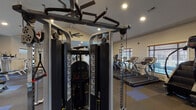 Located at 3305 10th St in Greenville, NC, this single-floor fitness center offers a comprehensive workout environment designed to accommodate a wide range of exercise needs. The space is defined by its vaulted ceilings, recessed lighting, and large ceiling fans that promote airflow throughout the facility.

The cardio zone lines one side of the floor with a row of Cybex treadmills and stair climbers, positioned near windows fitted with wooden blinds that allow natural light to filter in. A multi-station cable machine serves as a centerpiece for strength training, complemented by a variety of selectorized weight machines arranged across the open floor plan. Wall-to-wall mirrors on one end visually expand the space and assist with form during workouts.

A dedicated free weights area includes a full dumbbell rack alongside a flat bench, while a functional training corner features a power rack loaded with medicine balls, resistance tools, and a heavy bag for dynamic training. The flooring consists of durable rubberized tile in a neutral and blue checkered pattern, providing stability and cushioning throughout.

Glass double doors along one wall open to an outdoor area, enhancing the sense of openness. The overall layout is spacious and well-organized, making this fitness center a fully outfitted facility suitable for both cardio-focused and strength-based training routines.