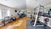 Located at 2726 Jacqueline Dr in Wilmington, Delaware, this single-floor fitness center is a well-appointed facility designed to support a full range of workout routines. The space features warm hardwood flooring throughout, complemented by neutral beige walls, recessed lighting, and vaulted ceilings that lend an open, airy feel. Natural light streams generously through multiple large windows fitted with white blinds, while a set of glass French doors with a transom window at one end provides additional brightness and a welcoming entry point.

The cardio zone is anchored by a row of Vision treadmills lined along one wall, alongside elliptical trainers and a stationary cycle, offering ample options for cardiovascular training. Wall-mounted televisions provide entertainment during workouts. The strength training area is equally comprehensive, featuring a Precor cable/weight machine, a Smith machine with bench, a multi-functional cable resistance unit, a dumbbell rack, weight plates, and a stability ball. Large wall mirrors in the strength zone enhance the sense of space and allow for proper form checks during exercise. Rubber floor mats protect the hardwood in high-use lifting areas. The overall layout is thoughtfully organized to separate cardio and strength zones, creating a functional, comfortable, and professional fitness environment.