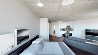 This single-floor space showcases a striking industrial-modern aesthetic, defined by its signature exposed concrete ceiling that lends the interior a raw yet refined character. The open-plan layout seamlessly integrates the living, sleeping, dining, and kitchen areas into one cohesive and functional environment.

The sleeping and living zone anchors the space with a neatly made bed, a light-toned area rug, and a comfortable sofa, creating a versatile area suited for both rest and relaxation. A framed artwork on the wall adds a touch of curated style to the space.

The kitchen is sleek and contemporary, featuring crisp white cabinetry, stainless steel appliances including a range, microwave, and refrigerator, and a clean countertop workspace. Adjacent to the kitchen, a compact dining area with a round table and dark chairs provides an intimate setting for meals.

Rich dark hardwood flooring runs throughout, offering a warm contrast to the cool concrete overhead and the white tones of the cabinetry and walls. A corridor visible in the background suggests additional private spaces, such as a bathroom. Recessed and flush-mounted ceiling lights ensure the space is well-lit. Overall, the design balances minimalist precision with functional comfort.