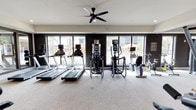 This fitness center offers a comprehensive, single-floor layout designed to support a wide range of workout routines. The main cardio area is anchored by a row of treadmills, elliptical trainers, and stationary and spin bikes, all positioned in front of expansive floor-to-ceiling windows that frame views of the surrounding outdoor environment, filling the space with natural light. Ceiling fans throughout ensure comfortable airflow during workouts.

The strength training zone features a full dumbbell rack with a broad weight range, weight benches, and multiple cable and resistance machines arranged along the perimeter, allowing for efficient use of the open floor space. Large mirrors amplify the sense of space and allow members to monitor their form.

A separate functional training studio provides a versatile open-carpeted area ideal for group exercise, stretching, or dynamic movement. This room is equipped with TRX suspension trainers mounted to a stylish shiplap wood accent wall, wall-mounted stability bars, and an assortment of stability balls. Natural light pours in through large windows, and framed artwork adds a welcoming touch to the neutral, contemporary décor. The overall design is modern and clean, with a neutral color palette, carpeted flooring, and recessed lighting that creates a motivating yet comfortable atmosphere.