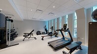 Located at 325 N 15th St in Philadelphia's vibrant Center City neighborhood, this fitness center offers a well-appointed and efficiently organized workout space across a single floor. The room is defined by a clean, modern aesthetic, with neutral gray walls, a drop tile ceiling with recessed lighting, and durable light-toned flooring throughout that gives the space a bright, polished feel.

A row of tall, floor-to-ceiling windows lines one wall, flooding the interior with natural light and providing views of the surrounding streetscape — creating an open, energizing atmosphere for exercise. The cardio section features treadmills, a stationary bike, an elliptical, and a rowing machine, all positioned to take advantage of the natural light. Strength training is well supported with a multi-functional cable machine, a full dumbbell rack displayed in front of a large wall mirror, adjustable benches, and a medicine ball storage station.

A dedicated mirror wall in the free weights area enhances the sense of space and supports form-focused training. The facility also includes a stability ball and yoga mat for stretching and functional fitness. Access to an adjacent room — likely a restroom or utility area — is visible through a glass-paneled door, adding to the facility's convenience. The overall ambiance is clean, modern, and purposeful.