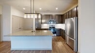 This single-story, one-bedroom, one-bathroom residence in Austin, TX offers a thoughtfully designed open-concept layout that maximizes functionality and flow. The kitchen serves as the centerpiece of the living area, showcasing rich dark-stained cabinetry extending to the upper walls, a full suite of stainless steel appliances including a refrigerator, range, microwave, and dishwasher, and a generous granite-topped island with an undermount sink. Pendant lighting with glass jar-style fixtures illuminates the island, adding a contemporary touch. White tile backsplash provides clean contrast against the dark cabinetry. Wood-style plank flooring runs throughout the main living areas, lending warmth and continuity. The living area is bright and open, featuring a ceiling fan, recessed lighting, and French doors that invite natural light and outdoor views. The bedroom is finished with plush carpet flooring and a large window offering exterior views. A walk-in closet with wire shelving provides practical storage. The bathroom features dark cabinetry with a granite-topped vanity, wood-style flooring, and direct access to a dedicated laundry area.