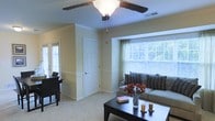 This single-floor residence centers around a welcoming living room that draws the eye with its large picture window dressed in horizontal blinds and sheer green curtains, allowing natural light to fill the space while offering views of lush greenery outside. The room features soft, neutral-toned walls accented by classic crown molding and a chair-rail detail that runs along the perimeter, adding a touch of architectural character. Plush wall-to-wall carpeting in a light beige tone contributes to the warm, comfortable ambiance throughout.

A ceiling fan with a built-in light fixture occupies the center of the room, providing both air circulation and ambient lighting. The layout accommodates a cozy seating area anchored by a sofa adorned with patterned pillows, paired with a dark wood coffee table that holds decorative candles and a floral arrangement, enhancing the inviting atmosphere.

Toward one end of the room, a dining or workspace area is partially visible, furnished with dark wood chairs. Multiple interior doors suggest access to additional rooms, and a secondary window near the entry area further brightens the space with natural light.