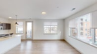 This single-floor residence features a clean, contemporary design throughout, characterized by crisp white walls, light hardwood-style flooring in the main living areas, and an abundance of natural light from multiple large windows fitted with blinds. The open-concept living area is generously proportioned, offering views of the surrounding neighborhood through a bank of windows, and includes direct access to a private balcony via a glass-paneled exterior door.

The kitchen is a standout feature, appointed with rich dark cabinetry, light stone-toned countertops, a white subway tile backsplash, and stainless steel appliances including a refrigerator, range, and microwave. A large kitchen island with pendant lighting and an integrated sink adds both functionality and style. The kitchen connects seamlessly to a hallway leading to the bedrooms.

The bedrooms are carpeted for comfort, each receiving natural light through well-sized windows. Two full bathrooms serve the unit; one features a soaking tub with subway tile surround, while the other offers a tiled walk-in shower. Both bathrooms include dark wood vanities with light countertops and hardwood-style flooring, maintaining a cohesive, modern aesthetic throughout.