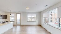 This single-floor residence features a clean, contemporary design throughout, characterized by crisp white walls, light hardwood-style flooring in the main living areas, and an abundance of natural light from multiple large windows fitted with blinds. The open-concept living area is generously proportioned, offering views of the surrounding neighborhood through a bank of windows, and includes direct access to a private balcony via a glass-paneled exterior door.

The kitchen is a standout feature, appointed with rich dark cabinetry, light stone-toned countertops, a white subway tile backsplash, and stainless steel appliances including a refrigerator, range, and microwave. A large kitchen island with pendant lighting and an integrated sink adds both functionality and style. The kitchen connects seamlessly to a hallway leading to the bedrooms.

The bedrooms are carpeted for comfort, each receiving natural light through well-sized windows. Two full bathrooms serve the unit; one features a soaking tub with subway tile surround, while the other offers a tiled walk-in shower. Both bathrooms include dark wood vanities with light countertops and hardwood-style flooring, maintaining a cohesive, modern aesthetic throughout.