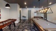 This single-floor space is designed as a contemporary game and lounge room, combining industrial aesthetics with comfortable, inviting elements. The most distinctive design feature is the polished epoxy flooring, which displays a dramatic marble-like pattern in deep grays and blacks, lending the space a bold, upscale character throughout.

The main area centers around a full-size pool table crafted in warm wood tones, illuminated overhead by a geometric, industrial-style pendant light fixture that serves as a focal point. Adjacent to the pool table stands a foosball table, providing additional entertainment options. A wall-mounted flat-screen TV is paired with a round bistro-height table and metal counter stools, creating a casual viewing and seating nook.

Along one wall, large windows fitted with horizontal blinds allow natural light to filter in, offering views of surrounding greenery. A plush accent chair positioned near the windows provides a relaxed seating option. The lounge area includes a cushioned sofa and wall-mounted décor, contributing warmth to the otherwise modern setting.

Dark-painted ceilings with recessed lighting and pendant fixtures enhance the moody, social atmosphere, making this space well-suited for recreation and gathering.