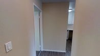 This single-story space is characterized by a straightforward, functional layout that connects its areas through a central hallway. The hallway features neutral beige walls with clean white trim and baseboards, complemented by dark wood-look flooring that runs throughout, adding a grounded, modern contrast to the light-toned walls.

To one side of the hallway, a doorway with white trim opens to an adjacent room equipped with a mirrored sliding door, suggesting a closet or storage space. The mirrored panel adds a reflective element that helps brighten the corridor. Flush-mount ceiling lighting provides consistent, even illumination throughout.

At the end of the hallway, the kitchen comes into view, featuring white upper cabinetry and a textured ceiling. The kitchen appears compact yet well-organized, with appliances visible and overhead lighting keeping the area well-lit. The consistent dark flooring flows seamlessly from the hallway into the kitchen, creating a cohesive visual connection between spaces.

Overall, the space presents a clean, neutral aesthetic with practical finishes, making it a functional and low-maintenance environment.