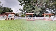 The grilling area is a well-maintained outdoor communal space designed for relaxed gatherings and outdoor dining. The centerpiece of the area is a covered wooden pavilion, providing shade and shelter over a classic picnic table — an ideal setup for outdoor meals regardless of weather conditions. A second pergola-style structure is positioned nearby, adding additional covered space for socializing or grilling activities.

The grounds are landscaped with a generous stretch of neatly kept green lawn, offering open space that complements the covered seating areas. A white picket fence borders the perimeter, clearly defining the space while contributing a clean, inviting aesthetic. Mature, leafy trees rise above the fence line, providing natural shade and a lush, canopied backdrop that enhances the sense of seclusion and tranquility.

The overall ambiance of the grilling area is casual and welcoming, suited for outdoor entertaining in a shared setting. The combination of structured pavilions, open lawn, natural greenery, and defined boundaries creates a functional yet pleasant environment. This space serves as a practical extension of the living area, encouraging residents to take advantage of outdoor communal living in a comfortable, well-organized setting.