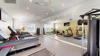 Located at 2500 McArthur Landing Cir in Fayetteville, NC, this dedicated fitness center offers a thoughtfully designed exercise room within a compact yet efficiently organized single-floor layout. The space is bright and welcoming, featuring white walls accented by warm sage-green paint, durable speckled flooring, and recessed lighting complemented by ceiling fans that promote air circulation during workouts. Floor-to-ceiling mirrors along one wall visually expand the room while allowing users to monitor their form throughout exercise sessions. Natural light streams in through multiple windows fitted with blinds, offering views of the surrounding area and contributing to the open, airy ambiance. The room is comprehensively equipped for a full-body workout, featuring treadmills, StairMaster step machines, a rowing machine, a stationary spin bike, and a multi-station cable strength-training machine. Additional equipment includes a dumbbell rack, free weights, exercise balls, resistance tools, and a flat-screen television mounted on the wall for entertainment during cardio sessions. The well-planned arrangement of equipment ensures ease of movement throughout the space, making it suitable for a variety of fitness routines from cardio and strength training to flexibility and functional fitness exercises.