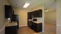 This single-story space showcases a well-appointed kitchen as its central feature, designed with dark espresso-toned cabinetry that extends from floor to ceiling, providing ample storage. The kitchen is equipped with a full suite of black appliances, including a refrigerator, electric range, built-in microwave, and dishwasher, all complemented by light-toned granite-style countertops that offer a striking contrast to the rich cabinetry. Overhead, a flush-mounted fluorescent light fixture provides bright, functional illumination throughout the cooking area. The kitchen flows seamlessly into adjoining living spaces, with an open sightline that enhances the sense of connectivity. Wood-style hard flooring runs through the kitchen and transitions into the surrounding areas, adding warmth and durability underfoot. Neutral, light-toned walls throughout maintain a clean and airy feel. White baseboards and trim provide crisp definition along the perimeter. A ceiling fan is visible in the adjacent room, hinting at comfortable, well-ventilated living spaces beyond the kitchen area.
