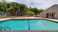 The Cedar Creek Circle Pool Area in Montgomery, Alabama is a well-defined outdoor recreational facility centered around a generously sized swimming pool with clear, blue-green water. The pool features classic brick coping along its edges and marked depth indicators, with entry points provided by built-in step ladders at multiple locations. A wide concrete deck surrounds the pool on all sides, offering ample space for lounging and outdoor furniture, including a metal table and chair set visible near the pool house. The facility is fully enclosed by a combination of chain-link and iron perimeter fencing, providing both security and visibility to the surrounding landscape. A dedicated pool house structure sits adjacent to the pool area, featuring a covered entrance with double doors, and appears to serve as an equipment and utility room. A life ring safety device is mounted on the exterior wall of the pool house. The backdrop features mature trees and lush greenery, creating a natural screen on one side, while the other side opens to views of the surrounding community and parking areas. The open-air setting allows for abundant natural light throughout the day, making this pool area a functional and inviting communal amenity for the Cedar Creek Circle community.