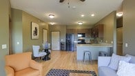This single-floor residence showcases an open-concept layout that effortlessly connects the living, dining, and kitchen areas into one cohesive and functional space. The living area is warm and inviting, featuring light hardwood flooring that extends throughout, a ceiling fan overhead, and a patterned area rug that anchors the seating arrangement. A cozy dining nook with a round glass-top table and upholstered accent chairs sits adjacent, adding an intimate touch to the open floor plan.

The kitchen is a standout feature, designed with rich medium-toned wood cabinetry, granite-style countertops, and black appliances including a French door refrigerator, electric range with overhead microwave, and a double-basin sink. A peninsula counter with industrial-style bar stools serves as both a functional prep space and a casual dining spot, creating a natural divide between the kitchen and living areas.

Recessed lighting, flush-mount ceiling fixtures, and a ceiling fan ensure the space is well-lit throughout. The soft, earthy green tones on the walls complement the warm flooring and cabinetry, lending the space a grounded, comfortable ambiance. A front entry door is visible from the main living area, reinforcing the practical, single-level flow of the home.