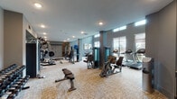This two-floor fitness facility in Frisco, TX is thoughtfully designed to accommodate a wide range of workout needs. The main floor boasts an expansive cardio and strength training area finished with neutral-toned carpet and accented by cool gray walls. It is well-equipped with multiple treadmills, elliptical trainers, cable machines, weight benches, and selectorized strength machines. Clerestory windows and large floor-to-ceiling glass panels allow generous natural light to flood the space, offering outdoor views that enhance the open, energizing atmosphere. Recessed lighting and wall-mounted televisions add comfort and convenience throughout.

A dedicated studio area on the lower level features full-length wall mirrors, decorative patterned sliding room dividers, and cork-style flooring. This zone houses cycling bikes, a rowing machine, dumbbells, and balance equipment, making it suitable for group or individual training sessions. Framed artwork and a refreshment station with a beverage cooler add a polished, welcoming touch.

A staircase with glass railings connects the two levels, while an entry vestibule with double doors provides a clean transition into the facility. Overall, the space combines functional fitness infrastructure with refined interior design elements for a professional and motivating workout environment.
