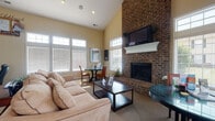 Located at 5222 Flower Valley Dr in Canal Winchester, Ohio, this single-floor community space is thoughtfully designed to serve both as a leasing center and a resident amenity hub. The main lounge area makes an immediate impression with its soaring vaulted ceilings, abundant natural light streaming through large multi-pane windows with transom accents, and a dramatic full-height brick fireplace serving as the room's focal point. A wall-mounted flat-screen television above a crisp white mantel complements the warm, inviting atmosphere, while carpeted flooring and neutral warm tones carry throughout the space. A cozy dining nook adjacent to the lounge provides a functional yet comfortable spot for meetings or casual gatherings. The leasing office area features display walls showcasing available floor plans and community photos, along with a dedicated workspace and ample natural light from oversized windows that offer pleasant outdoor views. A dedicated home theater room provides a standout amenity, equipped with a large projection screen, plush leather recliner seating, wall sconce lighting, and cinema-themed décor including decorative film reel wall accents. The overall ambiance of the space balances professionalism with residential warmth, making it both a practical leasing environment and an engaging showcase of community living.
