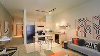 This single-floor space showcases a thoughtfully designed open-concept layout that seamlessly blends the kitchen and living areas into one cohesive environment. The kitchen is outfitted with light wood cabinetry, black appliances including a full-sized refrigerator and range, and a practical under-cabinet range hood. A compact kitchen island with a glass top and modern bar stools serves as a functional dining and gathering space, ideal for casual meals or entertaining. The countertops extend to a dedicated sink area, providing ample workspace throughout. Track lighting mounted on the ceiling offers adjustable, targeted illumination across the open floor plan, contributing to a warm and well-lit atmosphere. The living area flows naturally from the kitchen, anchored by a contemporary sofa accented with vibrant orange and gray throw pillows, complemented by a stylish floor lamp. Bold geometric artwork on the walls and a patterned area rug introduce pops of color and visual interest, giving the space a modern, curated aesthetic. The neutral-toned walls and light ceiling enhance the sense of openness, while the overall design strikes a balance between functionality and modern style.