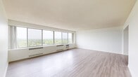 This well-appointed one-bedroom, one-bathroom unit is thoughtfully laid out across a single floor, offering a comfortable and efficient use of space. The living room is a standout feature, characterized by a wide bank of floor-to-ceiling windows that flood the room with natural light and frame panoramic views of a winding river and lush tree canopy below — a compelling focal point that lends the space an airy, elevated feel. Light-toned hardwood flooring runs throughout the main living areas, enhancing the sense of openness and brightness.

The galley-style kitchen is efficiently designed with white cabinetry, granite-look countertops, a stainless steel sink, and white appliances including a refrigerator and stove. An in-unit stacked washer and dryer adds everyday convenience. The kitchen connects seamlessly to the hallways, which guide residents through the unit's layout.

The bedroom continues the window-forward design, offering its own set of large windows with scenic outdoor views and ample natural light. Plush carpeting adds warmth underfoot, and a full wall of built-in louvered closets provides generous storage. The bathroom is clean and straightforward, finished with white tile flooring and a full tub-shower combination.