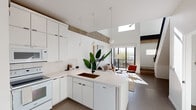 This two-story residence showcases a thoughtfully designed modern aesthetic throughout. The main level centers around an open-concept kitchen and living area, where soaring ceilings and exposed concrete beams create a striking industrial-contemporary contrast against the clean white walls and cabinetry. The kitchen features a large peninsula with a dark undermount sink, pendant lighting, full-height white cabinetry, tile backsplash, and a suite of white appliances. A skylight overhead floods the space with natural light, while large sliding glass doors connect the living area to an outdoor balcony, enhancing the sense of openness. The living area is anchored by the dramatic ceiling height and raw concrete beam detail, lending warmth and character to the bright, airy space. A second kitchen space on the lower level offers additional full cabinetry and white appliances, with a frosted glass entry door providing privacy. Upstairs, the bedrooms are carpeted and serene, featuring sliding closets, ceiling fans, and large windows that frame outdoor views. A lofted area near the staircase with a sleek glass and metal railing offers flexible workspace potential. Throughout, large-format tile flooring, crisp white walls, and an abundance of natural light define the cohesive and refined character of this multi-level home.