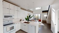 This two-story residence showcases a thoughtfully designed modern aesthetic throughout. The main level centers around an open-concept kitchen and living area, where soaring ceilings and exposed concrete beams create a striking industrial-contemporary contrast against the clean white walls and cabinetry. The kitchen features a large peninsula with a dark undermount sink, pendant lighting, full-height white cabinetry, tile backsplash, and a suite of white appliances. A skylight overhead floods the space with natural light, while large sliding glass doors connect the living area to an outdoor balcony, enhancing the sense of openness. The living area is anchored by the dramatic ceiling height and raw concrete beam detail, lending warmth and character to the bright, airy space. A second kitchen space on the lower level offers additional full cabinetry and white appliances, with a frosted glass entry door providing privacy. Upstairs, the bedrooms are carpeted and serene, featuring sliding closets, ceiling fans, and large windows that frame outdoor views. A lofted area near the staircase with a sleek glass and metal railing offers flexible workspace potential. Throughout, large-format tile flooring, crisp white walls, and an abundance of natural light define the cohesive and refined character of this multi-level home.