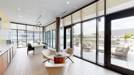 This contemporary amenity lounge is designed with an open, airy feel anchored by expansive floor-to-ceiling glass walls and sliding doors that flood the interior with natural light and provide seamless access to a furnished outdoor terrace. The light wood-plank flooring runs throughout, complementing the clean, neutral palette of the main gathering area, which features modern curved lounge chairs, a sculptural wood side table, and sleek pendant lighting suspended from a recessed ceiling.

A built-in kitchenette lines one wall, finished with warm wood cabinetry, a white quartz countertop, an integrated sink, a compact dishwasher, and a wall-mounted flat-screen television set against a textured golden tile backsplash — ideal for casual entertaining or events.

A separate lounge zone makes a bold artistic statement with a vibrant, large-scale tropical mural spanning the full wall, accented by woven hanging pod chairs and a sculptural globe chandelier. This area opens directly to the terrace through glass-paned French doors, reinforcing the indoor-outdoor connection.

Steel-framed glass partitions divide the space while maintaining visual openness throughout. The terrace beyond offers additional seating and sweeping views of the surrounding urban landscape, making this lounge a versatile and visually dynamic communal space.