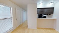 This single-floor space showcases a clean, minimalist design with light-toned hardwood flooring that runs throughout, contributing to a bright and cohesive aesthetic. The layout is open and functional, with a structural column that subtly divides the main living area from the kitchen zone without fully enclosing either space.

The kitchen is positioned to the right and features a pass-through counter with a dark upper cabinet overhead, creating a visual contrast against the white walls. Black appliances, including what appears to be a refrigerator and range, are visible from the main area, and a sink is integrated into the countertop. The dark cabinetry provides a bold accent within an otherwise light interior.

A hallway extends toward the back of the space, lined with white doors — one appearing to be an entry or utility door and another a closet with standard bifold or panel-style doors. The white walls and flush ceiling-mounted light fixture keep the space feeling clean and uniform. Baseboards throughout are bright white, reinforcing the tidy, well-maintained character of the unit. Overall, the space offers a practical and uncluttered environment suited for everyday living.
