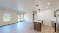 This single-floor space showcases a clean, contemporary design with a cohesive neutral palette throughout. The open-concept layout connects the kitchen and living area seamlessly, creating a bright and functional environment. Light wood-look luxury vinyl plank flooring runs throughout, contributing to the space's airy, modern aesthetic.

The kitchen is well-appointed with rich, medium-toned wood cabinetry featuring sleek hardware, light marble-style countertops, and a full suite of stainless steel appliances, including a Whirlpool refrigerator, electric range, and over-the-range microwave. A peninsula island with an integrated double sink and matching countertop offers additional workspace and casual seating. Pendant lighting with exposed Edison-style bulbs hangs above, adding a contemporary touch.

The living area is generously proportioned with white walls and recessed lighting. A glass-panel patio door fitted with a black barn-style hardware rail floods the space with natural light and provides outdoor access, while an additional window enhances ventilation and views.

The bathroom features a dual-sink vanity with matching wood cabinetry, marble-style countertops, and well-lit mirrors, maintaining the design consistency found throughout the rest of the space. Black hardware accents on doors and fixtures provide a refined finishing detail.