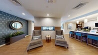 This thoughtfully designed single-floor amenity space offers a generous and versatile layout that blends social, recreational, and functional areas seamlessly. Warm wood-tone flooring flows throughout, creating a cohesive and inviting atmosphere across all zones.

The lounge area serves as the welcoming heart of the space, featuring a bold geometric-patterned accent wall in deep navy and gold, a built-in media wall with open wood shelving and a large flat-screen TV, and plush seating including high-back armchairs and a circular accent rug. Wall sconces and recessed lighting provide a warm, layered ambiance.

A central divider with a pass-through window and a built-in bench seat with decorative pillows elegantly separates the lounge from the entry corridor, which leads to a secondary seating area with natural light from large exterior-facing windows and a round mirror accent.

The kitchen features rich walnut cabinetry, open display shelves, a white quartz island with bar-height seating in teal upholstered chairs, pendant lighting, a microwave, coffee station, and a wall-mounted TV — ideal for casual gatherings.

The dining area offers a long communal table surrounded by blue velvet chairs, bathed in natural light through floor-to-ceiling windows with outdoor views.

The game room is accessed through a wide wood-framed opening and includes a full-size pool table, soft carpet tile flooring, lounge seating, a wall-mounted TV, and vibrant artwork, making it a dynamic recreation destination.