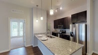 This single-floor residence offers a thoughtfully designed open-concept layout that seamlessly connects the main living areas. The kitchen serves as a focal point, featuring striking granite countertops on a large island with an undermount sink, rich dark espresso cabinetry, stainless steel appliances, and dramatic pendant lighting that draws the eye upward toward the soaring ceilings. Track lighting adds additional illumination throughout the cooking space.

The living area is generously proportioned, finished with warm hardwood-style flooring and a ceiling fan overhead. Natural light pours in through multiple windows and a set of French doors that open to outdoor views, creating a bright and airy atmosphere. The layout flows conveniently toward additional rooms, including well-sized bedrooms carpeted for comfort, each benefiting from abundant natural light through large windows with blinds.

The primary bathroom features a glass-enclosed walk-in shower, dark vanity cabinetry with a white countertop, and clean tile flooring. A secondary bathroom offers a tub-shower combination, dark cabinetry, a large vanity mirror, and wood-style flooring. Completing the unit is a dedicated laundry room equipped with a full-sized top-loading washer and dryer alongside built-in wire shelving for additional storage.