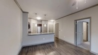 This single-floor residence showcases a thoughtfully designed open-concept layout that seamlessly connects the kitchen and living areas. The kitchen is anchored by a substantial peninsula with a dark granite-style countertop, offering both additional prep space and casual seating. Pendant lighting hangs above the peninsula, providing focused illumination and a modern aesthetic touch. Stainless steel appliances, including a full-size refrigerator, complement the dark cabinetry visible in the cooking area, creating a contemporary contrast against the light gray walls.

The flooring throughout is a rich, dark hardwood-style material that adds warmth and visual depth to the space. Neutral gray walls and white ceilings contribute to a clean, cohesive palette. The open living area offers generous space for furniture arrangement, and crown molding details along the ceiling add a refined architectural element.

Multiple doors visible along the hallway suggest additional rooms beyond the main living space. The layout promotes an efficient and connected flow, making the space feel airy and well-organized, while the consistent finishes throughout lend a polished, move-in-ready appearance.