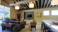 This single-floor space blends modern industrial design with comfortable, welcoming interiors. The most striking architectural feature is the exposed beam ceiling, which is complemented by visible ductwork running along its length, lending the space an open, loft-like character. Pendant drum lights and a ceiling fan are suspended from the beams, providing both ambient illumination and air circulation throughout the area.

The main lounge area is anchored by rich brown leather sofas and accent chairs arranged around a central coffee table, creating an inviting gathering zone. A recessed accent wall, finished in a deep olive-green hue, frames a wall-mounted flat-screen television, serving as a natural focal point for the room. Neutral-toned carpet underfoot adds warmth and comfort to the space.

Colorful abstract artwork adorns the walls, adding pops of visual interest against the warm beige tones. Decorative greenery and tall sculptural arrangements contribute a natural, organic touch to the overall aesthetic. To one side, a counter-height bar table with sleek stools offers an additional seating area, ideal for casual use. Natural light filters in through a window, brightening the space and enhancing its open, airy feel. Overall, the space exudes a contemporary yet relaxed ambiance.