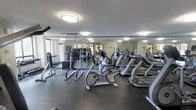 This fitness center offers a practical and well-organized space for residents or members to pursue their health and wellness goals. The room is outfitted with a solid selection of cardio equipment, including multiple treadmills and a stationary recumbent bike, positioned to allow comfortable spacing between machines. A dedicated strength training area features a cable/pull-up tower and a dumbbell rack, providing options for a full-body workout without the need to leave the facility.

Large mirrors line the walls, visually expanding the space and allowing users to monitor their form during exercise. The dark rubber flooring provides durability and cushioning underfoot, making it well-suited for high-impact activity. Overhead flush-mount lighting ensures the room is consistently and evenly illuminated throughout.

A textured ceiling adds a classic finish to the space, while a window along one wall introduces a degree of natural light and a connection to the outdoors. The overall layout is efficient and uncluttered, offering a clean and functional environment that accommodates both cardio and strength training needs within a single, cohesive space.