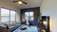 This single-floor model space showcases a contemporary interior design aesthetic that balances comfort with modern style. The living area serves as the focal point of the space, featuring a striking dark charcoal accent wall that adds depth and contrast against the lighter surrounding walls. Large windows dressed with horizontal blinds allow generous natural light to filter into the room, creating a warm and inviting ambiance throughout the day.

A ceiling fan with an elegant, decorative fixture provides both functionality and visual interest overhead. The living area is furnished with a light grey sofa accented by yellow and patterned throw pillows, complemented by a round wooden coffee table atop a classic patterned area rug. A cozy accent chair with a throw blanket adds an additional seating option near the windows.

Rich, dark hardwood-style flooring runs throughout, grounding the space and offering a pleasing contrast to the lighter furnishings. A substantial wooden media console along one wall provides ample storage and display space, enhancing the room's warm, layered feel. Thoughtful decorative touches, including floor lamps, greenery, and curated accessories, contribute to an overall atmosphere that is both stylish and livable. This model space effectively demonstrates the design potential of the floor plan.