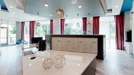 This single-floor residential lobby and mail center is thoughtfully designed to blend functionality with contemporary style. The entryway greets visitors with light wood-look flooring, bold geometric wall art in pink, blue, and teal tones, and a welcoming mural that adds a lively, artistic character to the space. Large windows along the entry allow natural light to filter in, creating an open and inviting atmosphere.

The mail and package center serves as the hub of the space. Individual mailboxes are neatly arranged along the walls, complemented by a large bank of automated package lockers that provide secure, convenient parcel retrieval. A dedicated workstation with a desktop computer is available for resident use. A wall-mounted display screen adds a modern, connected touch to the area.

Overhead, faceted geometric pendant lights in a warm metallic finish hang from the ceiling, adding a sophisticated decorative element. In the adjacent area, a teal-painted ceiling with deep crimson drapery panels frames large windows overlooking lush greenery, lending the space a bold, design-forward aesthetic. A generous quartz-topped counter provides a practical surface for sorting mail. Vibrant artwork, including a striking panther painting, further enriches the space's dynamic and modern character throughout.