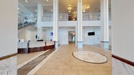 Located at 2500 Edwards Dr in Fort Myers, Florida, this impressive lobby space makes a striking first impression with its soaring double-height ceilings and expansive, open layout. The main entrance is framed by floor-to-ceiling glass walls and automatic sliding doors, flooding the interior with natural light and offering clear views of the surrounding waterfront and palm tree-lined exterior.

The flooring throughout is a combination of large-format beige tile and polished marble accents, including a distinctive circular marble medallion inlaid at the center of the main hall. Tall, marble-clad columns rise dramatically to the upper mezzanine level, which is lined with a sleek metal railing and visible from the ground floor, reinforcing the sense of vertical grandeur.

A curved reception desk with a rich wood veneer base and white countertop serves as the central service point, accompanied by wall-mounted displays. Adjacent to the entrance is a comfortable lounge area outfitted with plush white seating and a wood-framed coffee table, positioned to take full advantage of the panoramic outdoor views. A floating open-tread staircase leads to the upper level, with a workstation positioned beneath it. Warm wood column cladding, recessed and pendant lighting, and integrated LED column lighting complete the polished, contemporary aesthetic of this welcoming commercial lobby.