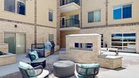 This thoughtfully designed outdoor courtyard is nestled within the interior of a contemporary multi-story building, creating a sheltered and inviting communal environment. The architecture features a warm combination of light-toned brick and neutral panel siding, with balconies overlooking the courtyard from upper floors, reinforcing a sense of verticality and enclosure.

At the heart of the courtyard stands a substantial double-sided brick fireplace, serving as both a functional focal point and a natural divider within the space. Adjacent to it, a dedicated outdoor kitchen area features a large stainless steel gas grill set within a brick surround, ideal for gatherings and outdoor entertaining.

The courtyard is furnished with modern rope-woven lounge chairs and ottomans accented with teal and white cushions, arranged in multiple conversational groupings across a smooth concrete patio surface. String lights are strung overhead, adding a warm, ambient glow that enhances the space during evening hours.

Raised planter beds with low-growing shrubs and greenery are integrated throughout, softening the hardscape and introducing natural elements. Additional seating nooks are tucked along the building perimeter, offering quieter retreats. Large glass entry doors provide seamless interior access, while open sky above floods the courtyard with natural light throughout the day.