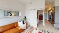 This contemporary single-floor residence offers a thoughtfully designed layout that maximizes both comfort and style. The living and dining area is the heart of the space, anchored by expansive floor-to-ceiling windows that frame sweeping urban skyline views, flooding the interior with natural light. Light hardwood-style flooring, colorful geometric area rugs, and carefully curated furnishings — including a cognac leather sofa and terracotta dining chairs — lend a warm, modern aesthetic. A mid-century credenza and bold abstract wall art add character to the living area. The kitchen is outfitted with white cabinetry, clean quartz countertops, stainless steel appliances, and a striking chevron-patterned tile backsplash, creating a functional yet visually appealing culinary space. The bathroom features large-format tile flooring, a wood-toned vanity with ample drawer storage, a white quartz countertop, a wide mirror, and a glass-enclosed shower. A standout architectural detail is the sliding barn door that connects the living area to the private bedroom, offering both a stylish design element and flexible space separation. Throughout, the crisp white walls and neutral palette create a bright, airy ambiance well-suited to contemporary urban living.