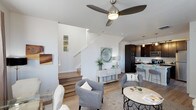 This well-appointed two-story home showcases a thoughtfully designed open-concept main floor that connects the living and kitchen areas in a seamless, functional layout. The living area is anchored by light hardwood flooring, a ceiling fan with a sleek brushed nickel finish, and crisp white walls that create a clean, airy atmosphere. A staircase with white risers and a light wood tread leads to the upper level, serving as an architectural feature that adds visual interest to the space. The kitchen is finished with rich dark cabinetry, providing a striking contrast to the bright surroundings. A pendant light hangs above a raised breakfast bar with seating, making it an ideal spot for casual dining or entertaining. Stainless steel appliances complete the kitchen's modern aesthetic. Throughout the main floor, natural light fills the space, enhancing the warm tones of the hardwood floors and complementing the neutral color palette. The overall ambiance is contemporary and welcoming, with a balance of light and dark tones that gives the home a refined yet comfortable character. The two-floor layout offers distinct living zones, providing both communal and private areas suited to a variety of lifestyles.