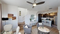 This well-appointed two-story home showcases a thoughtfully designed open-concept main floor that connects the living and kitchen areas in a seamless, functional layout. The living area is anchored by light hardwood flooring, a ceiling fan with a sleek brushed nickel finish, and crisp white walls that create a clean, airy atmosphere. A staircase with white risers and a light wood tread leads to the upper level, serving as an architectural feature that adds visual interest to the space. The kitchen is finished with rich dark cabinetry, providing a striking contrast to the bright surroundings. A pendant light hangs above a raised breakfast bar with seating, making it an ideal spot for casual dining or entertaining. Stainless steel appliances complete the kitchen's modern aesthetic. Throughout the main floor, natural light fills the space, enhancing the warm tones of the hardwood floors and complementing the neutral color palette. The overall ambiance is contemporary and welcoming, with a balance of light and dark tones that gives the home a refined yet comfortable character. The two-floor layout offers distinct living zones, providing both communal and private areas suited to a variety of lifestyles.