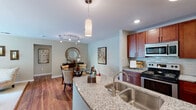 This single-floor residence at 7012 Gentle Shade Rd in Columbia, Maryland offers a thoughtfully designed layout that balances comfort and functionality. The open-concept kitchen is outfitted with rich cherry wood cabinetry extending to the ceiling, granite countertops, a double-basin stainless steel sink, subway tile backsplash, and stainless steel appliances including a range, microwave, and refrigerator. A granite-topped island provides additional prep space and flows seamlessly into the dining area, which features warm hardwood flooring and stylish track lighting overhead. The living room is carpeted and filled with natural light from a large sliding glass door that opens to an outdoor view. Built-in shelving adds practical storage and display space. The primary bedroom offers plush carpeting and a closet with built-in shelving, while a secondary room with similar carpeting and a closet serves well as a bedroom or home office. The full bathroom includes a granite vanity with cherry wood cabinetry, tile flooring, and a tub-shower combination. Throughout, a neutral color palette of soft grays and warm beiges creates a cohesive, inviting atmosphere across every room.