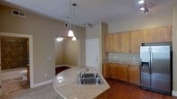 This single-floor residence offers a well-designed open-concept layout that seamlessly connects the kitchen and living areas. The kitchen is a standout feature, boasting warm honey-toned wood cabinetry that provides ample storage space, complemented by granite countertops that add a touch of elegance. A curved kitchen island with a double stainless steel sink serves as a functional centerpiece, ideal for both meal preparation and casual dining. Pendant lighting hangs above the island, adding character and focused illumination to the space. The kitchen flows naturally into the adjacent living area, which is finished with rich hardwood flooring and features a ceiling fan for year-round comfort. Neutral-toned walls throughout create a warm, inviting ambiance that lends itself well to a variety of interior styles. The entry door is conveniently accessible from the main living space, enhancing the practical layout of the residence. Recessed and surface-mounted lighting fixtures provide consistent, well-distributed light across the interior. The overall design balances functionality with a comfortable, approachable aesthetic, making this a practical and attractive living space.