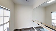 This yoga studio is a purpose-built, single-floor space thoughtfully designed to support movement, mindfulness, and physical practice. The studio features a clean, minimalist aesthetic defined by smooth, white walls and bright, neutral tones that create a calm and focused atmosphere ideal for yoga and fitness activities.

A standout feature of the studio is the large floor-to-ceiling wall mirror, which spans a significant portion of one wall. This mirror not only enhances the sense of openness in the room but also allows practitioners to monitor their form and alignment during sessions. Complementing the mirror is a wooden ballet barre mounted at an accessible height along the same wall, offering support for stretching, balance work, and barre-style exercises.

The flooring consists of patterned carpet tiles in a dark, geometric design, providing a practical and comfortable surface underfoot. Overhead, a ceiling vent ensures proper air circulation throughout the space, contributing to a comfortable environment during active sessions.

The overall ambiance of the studio is serene and uncluttered, with ample open floor space that can accommodate a variety of yoga styles, stretching routines, or small group classes. The combination of reflective surfaces, natural wood elements, and a neutral color palette makes this studio a versatile and inviting environment for practitioners of all levels.