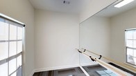 This yoga studio is a purpose-built, single-floor space thoughtfully designed to support movement, mindfulness, and physical practice. The studio features a clean, minimalist aesthetic defined by smooth, white walls and bright, neutral tones that create a calm and focused atmosphere ideal for yoga and fitness activities.

A standout feature of the studio is the large floor-to-ceiling wall mirror, which spans a significant portion of one wall. This mirror not only enhances the sense of openness in the room but also allows practitioners to monitor their form and alignment during sessions. Complementing the mirror is a wooden ballet barre mounted at an accessible height along the same wall, offering support for stretching, balance work, and barre-style exercises.

The flooring consists of patterned carpet tiles in a dark, geometric design, providing a practical and comfortable surface underfoot. Overhead, a ceiling vent ensures proper air circulation throughout the space, contributing to a comfortable environment during active sessions.

The overall ambiance of the studio is serene and uncluttered, with ample open floor space that can accommodate a variety of yoga styles, stretching routines, or small group classes. The combination of reflective surfaces, natural wood elements, and a neutral color palette makes this studio a versatile and inviting environment for practitioners of all levels.