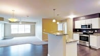 This single-floor residence showcases an open-concept design that seamlessly connects the kitchen, dining, and living areas. The kitchen is a standout feature, equipped with dark granite countertops that extend across both a peninsula and additional counter space, providing ample room for meal preparation. White cabinetry adds a clean, classic contrast to the rich countertop surfaces, while two elegant pendant lights hang above the peninsula, casting warm, focused illumination. A second sink is visible along the back kitchen wall, enhancing the space's functionality.

The accent walls — one in a warm golden-yellow tone and another in deep eggplant purple — add visual depth and character throughout the open layout. Transitioning from the kitchen, the hardwood-style flooring gives way to a carpeted living area, offering a comfortable and defined space for relaxation. A window along the far wall invites natural light into the living room, brightening the neutral gray tones of the walls and carpet. The overall ambiance is modern and inviting, with thoughtful design details that balance practicality with aesthetic appeal.
