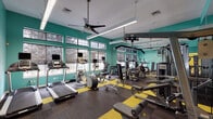 This fitness center is a comprehensively outfitted facility designed to support a wide range of workout routines. The space is divided into distinct functional zones, each thoughtfully appointed to maximize usability and comfort.

The main workout floor is the centerpiece of the facility, featuring bold teal walls accented by vivid yellow floor tiles set against dark carpet. The room is lined with large, grid-paned windows that allow an abundance of natural light to fill the space, while ceiling fans and suspended linear lighting ensure a comfortable environment. The area is equipped with a full suite of cardio machines — including treadmills, ellipticals, and stair climbers — as well as strength training stations, cable machines, and a dumbbell rack. Full-length mirrors along one wall expand the sense of space and allow users to monitor their form. Metal lockers are conveniently integrated into the room.

A separate studio area provides warm wood-look flooring, mirrored walls, and large windows, making it ideal for spinning, yoga, or stretching sessions. A spinning bike and dumbbell rack are present, and the open floor plan accommodates group fitness activities. Colorful exercise balls and accessories are neatly stored nearby.

Together, the zones create an energetic and well-organized fitness environment suited for both individual training and group exercise.